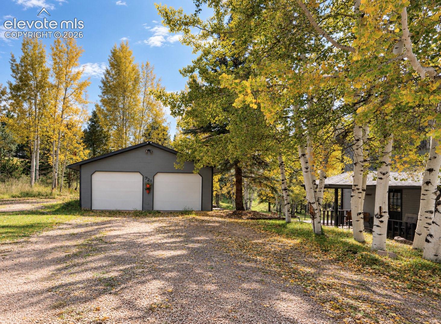 Detached two-car garage with white garage doors and gray siding, gravel driveway, and mature trees