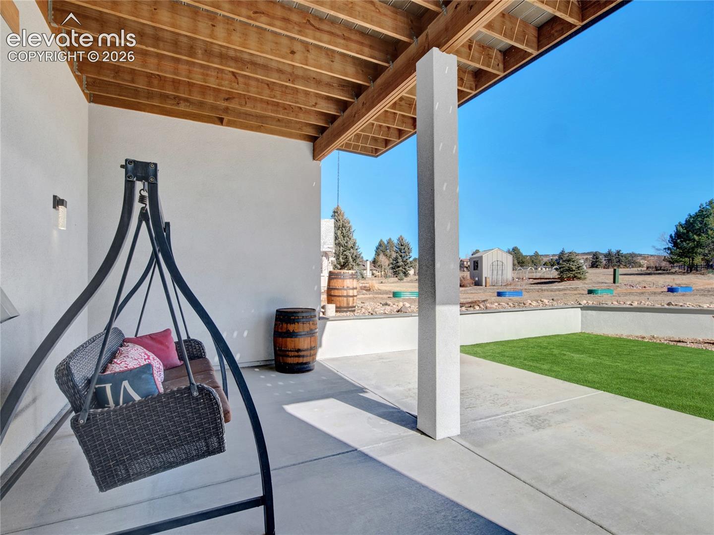 Covered patio with concrete flooring, exposed wood ceiling beams, a white stucco wall, and a fixed outdoor light fixture