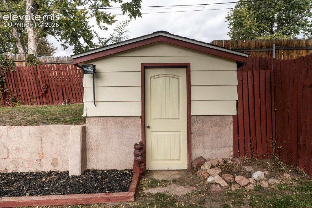 Detached storage structure featuring horizontal lap siding, a textured masonry foundation, a singular arched-panel door, and an exterior security light