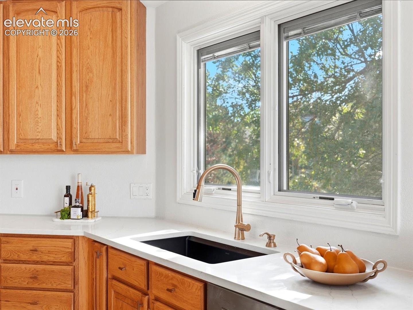 Natural wood-finish cabinetry, white solid-surface countertops, undermount rectangular sink, and high-arc metallic pull-down faucet beneath a dual-pane window