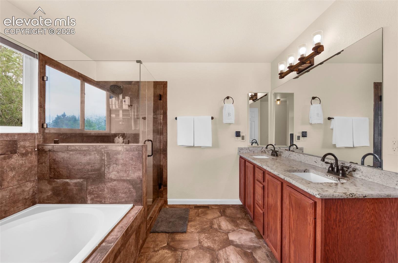 Bathroom featuring a large soaking tub with a tiled surround, a glass-enclosed shower, a double vanity with granite countertops, wood-finish cabinetry, and stone-look flooring
