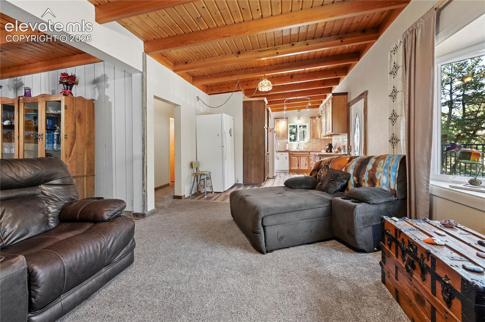 Vaulted wood-paneled ceiling with exposed beams, carpeted flooring, kitchen with wood cabinetry, and a window with outdoor scenery
