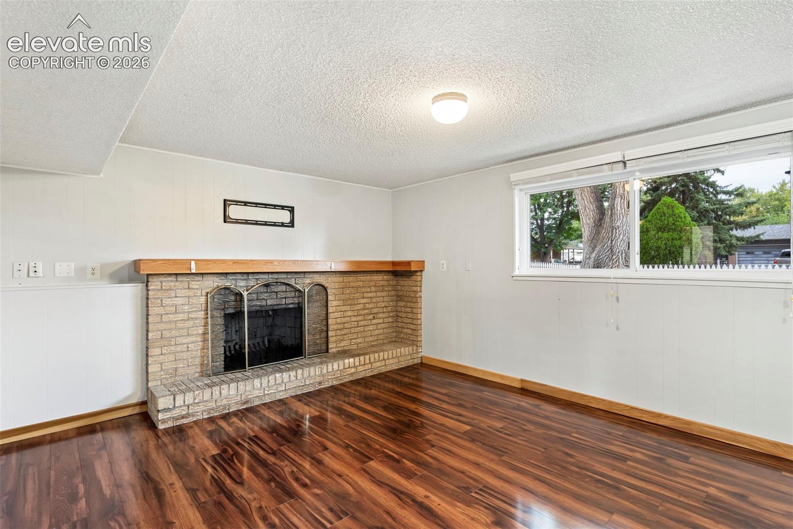 Brick fireplace with a wood mantel, wood-finish flooring, and a large window
