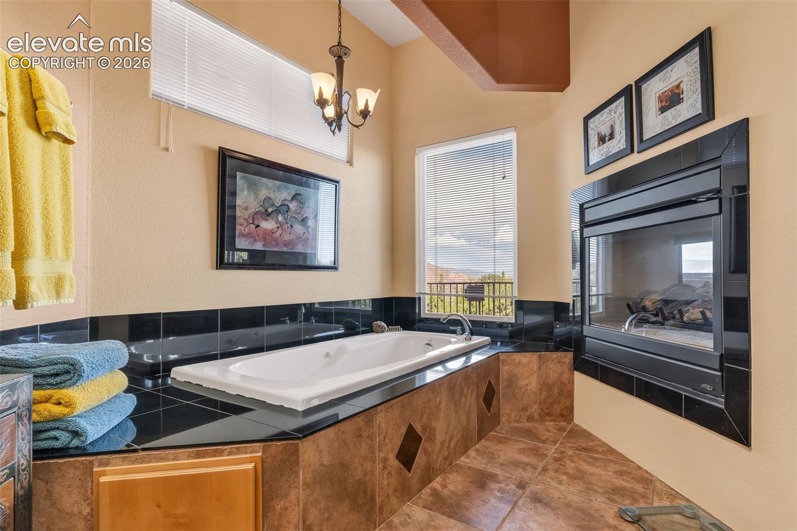 Soaking tub with black tile surround, built-in gas fireplace with black trim, tile flooring, and multi-light hanging chandelier