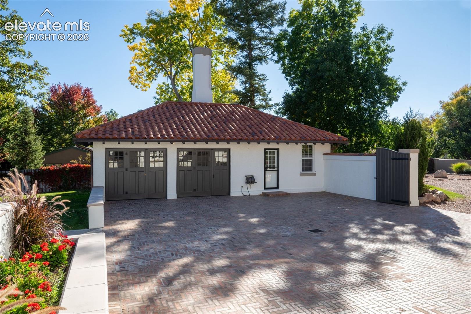 Detached two-car garage featuring a terracotta tile roof, white stucco exterior, prominent central chimney, and a herringbone-patterned brick paver driveway