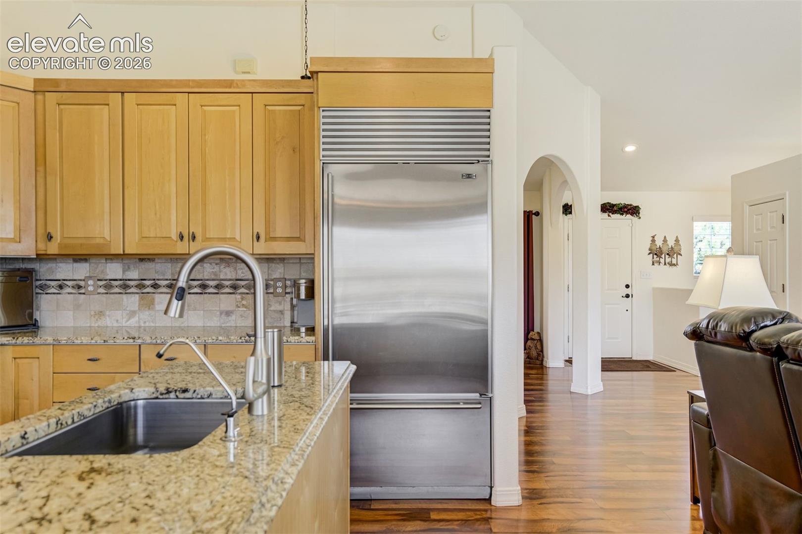 Natural wood cabinetry featuring a granite countertop, stainless steel refrigerator with integrated cabinetry surround, stone tile backsplash, stainless steel sink with a high-arch faucet, and arched architectural detailing