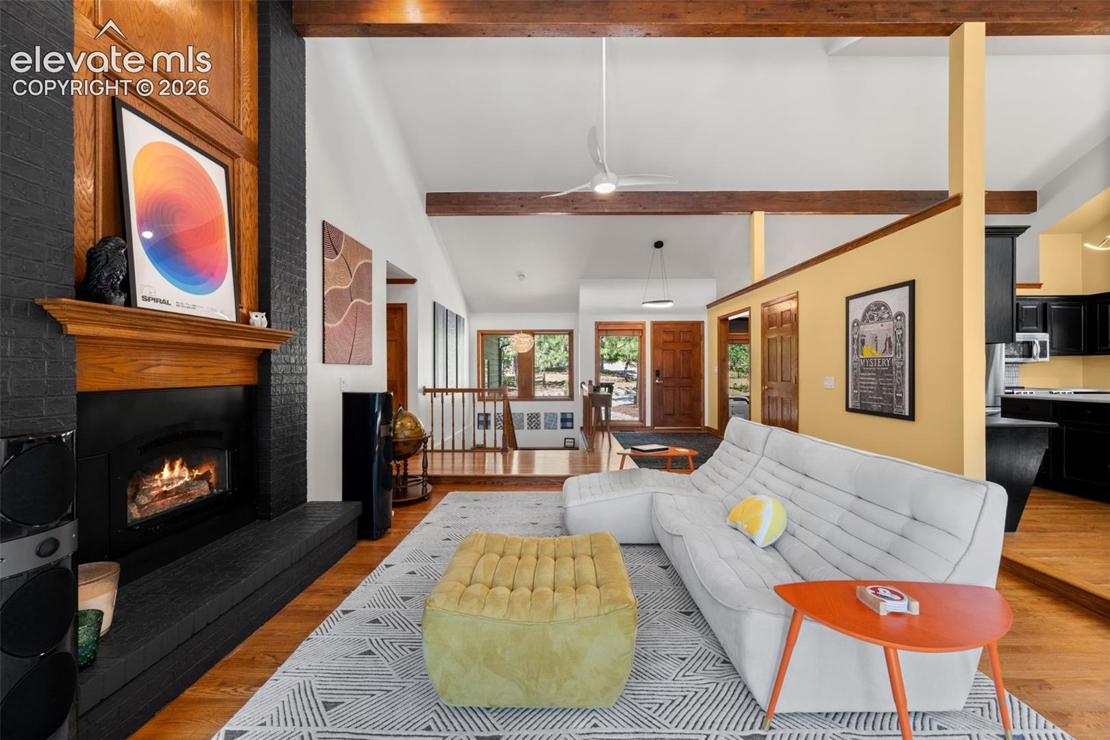 Spacious living area featuring a black brick fireplace with a wood mantel, exposed ceiling beams, wood-finish flooring, and a partial wall separating a kitchen area with black cabinetry