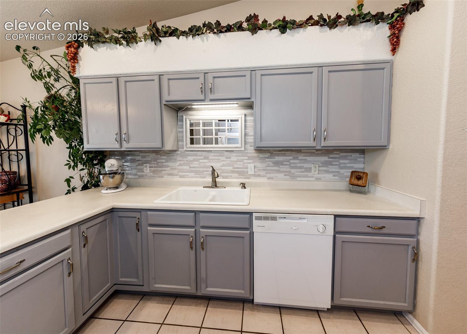 Kitchen featuring painted grey cabinetry, a multi-tone linear tile backsplash, integrated white double-basin sink, and a built-in dishwasher