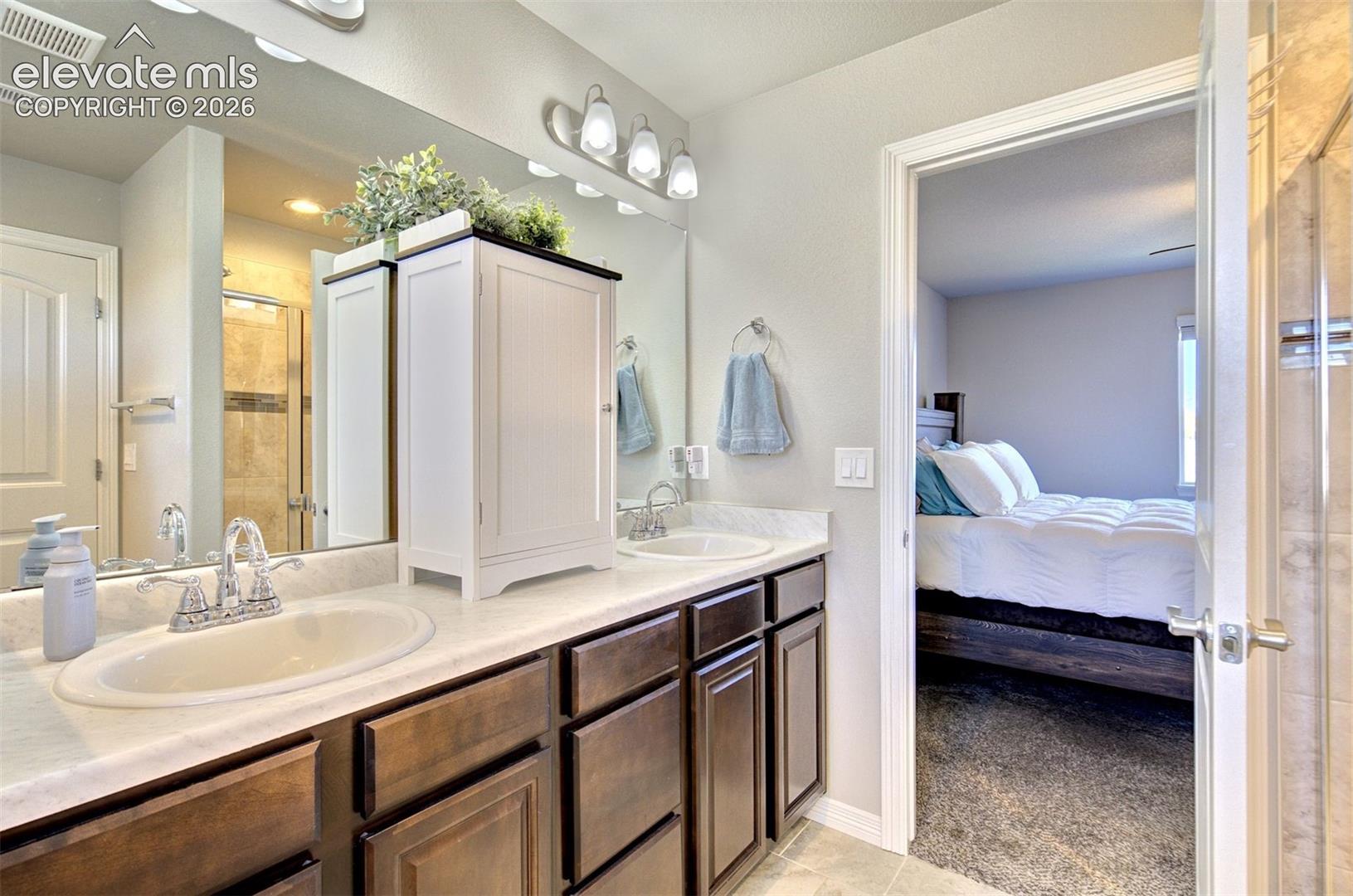 Bathroom featuring a dual-sink vanity with light-toned stone-finish countertop, dark wood-finish cabinetry, full-width mirror, and tri-light fixture