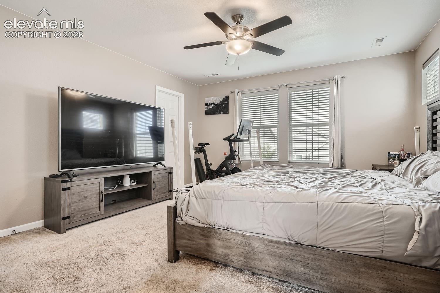 Neutral-toned room featuring plush wall-to-wall carpeting, dual windows with horizontal blinds, and a central ceiling fan with integrated lighting