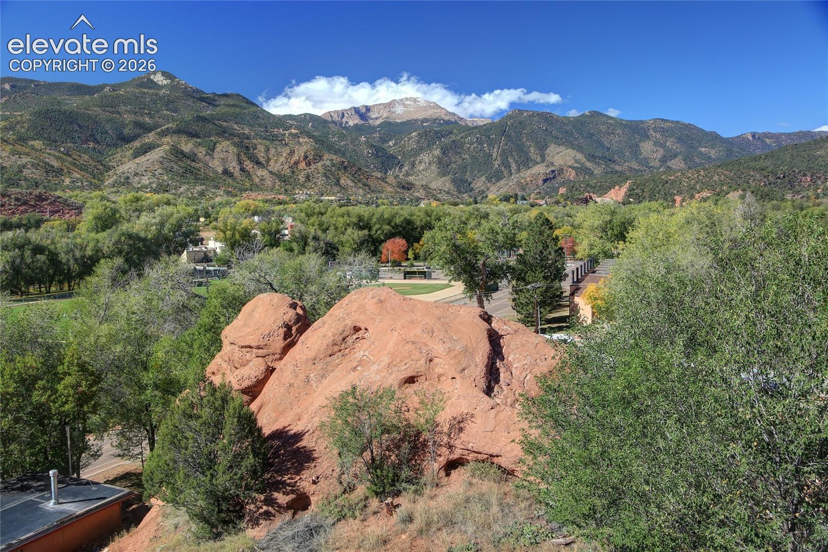 Expansive mountain landscape featuring prominent red rock formations, mature deciduous trees, and coniferous evergreens