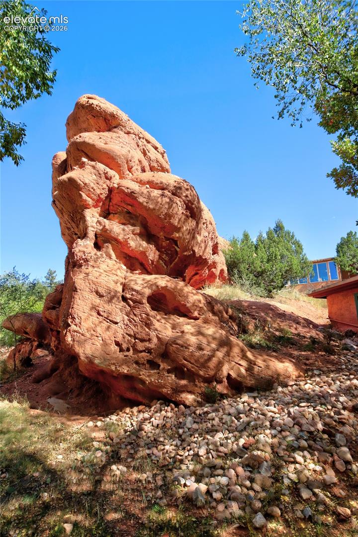 Prominent natural red rock formation set against a landscape of mature trees and stone ground cover
