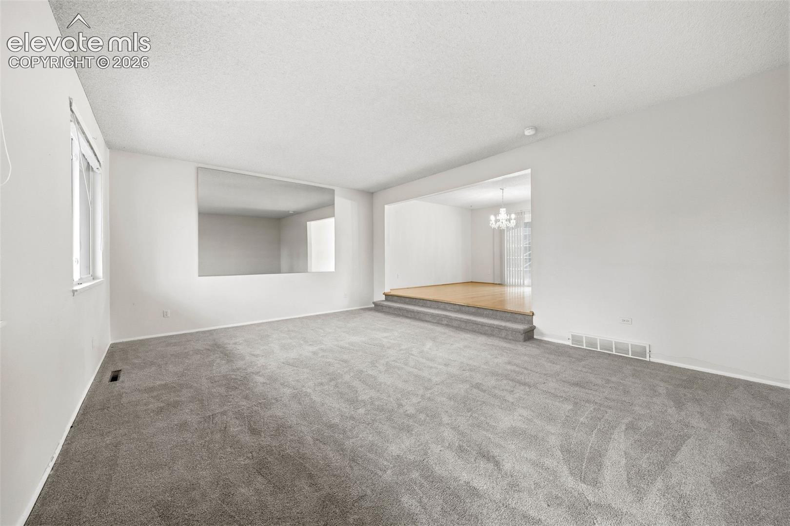 Open-concept interior featuring plush wall-to-wall carpeting, an inset wall mirror, a raised platform with wood-finish flooring, and a multi-arm chandelier