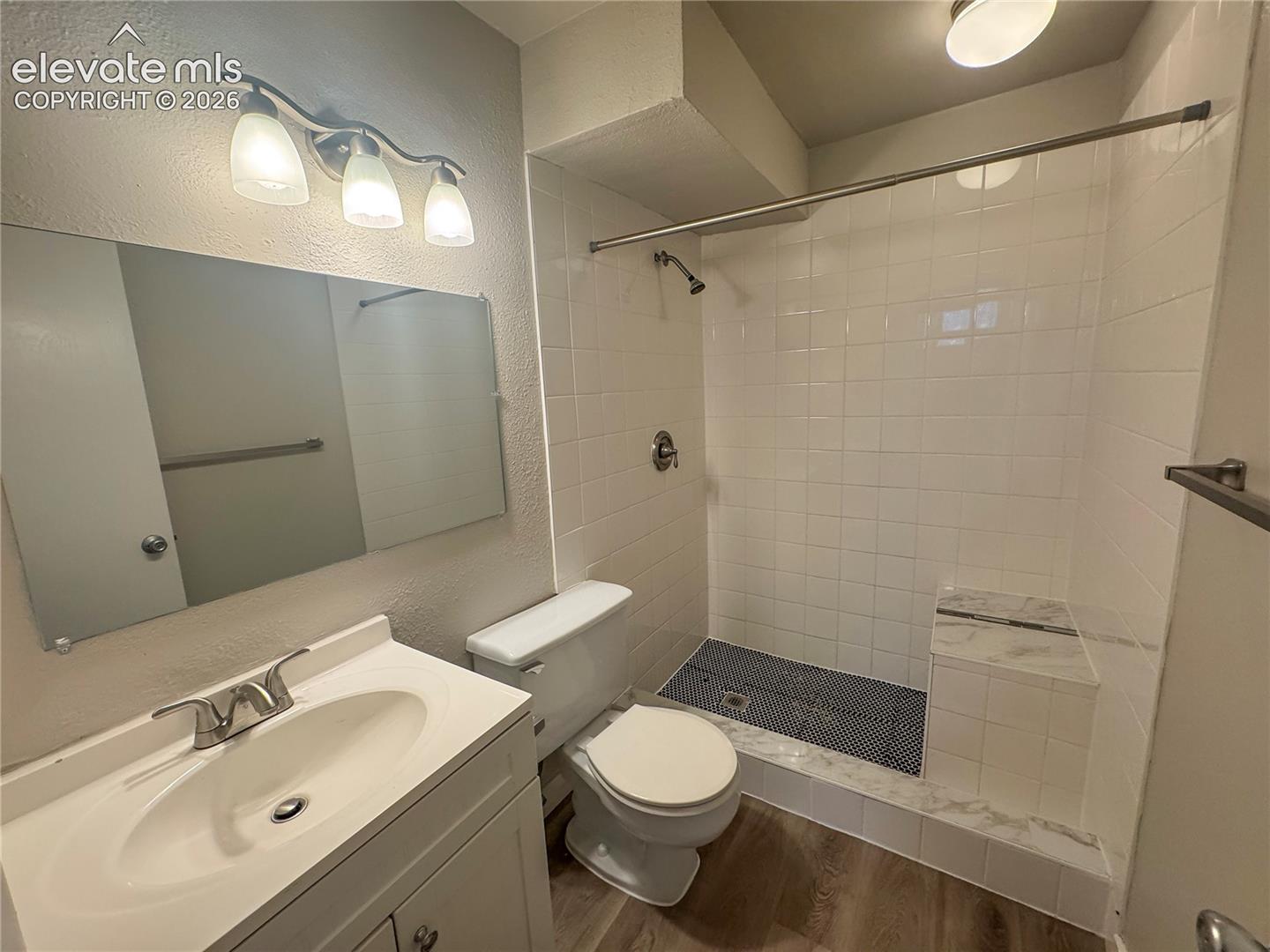 Bathroom featuring wood-finish flooring, a vanity with an integrated sink, a frameless wall mirror, and a walk-in shower with tiled walls and a built-in bench