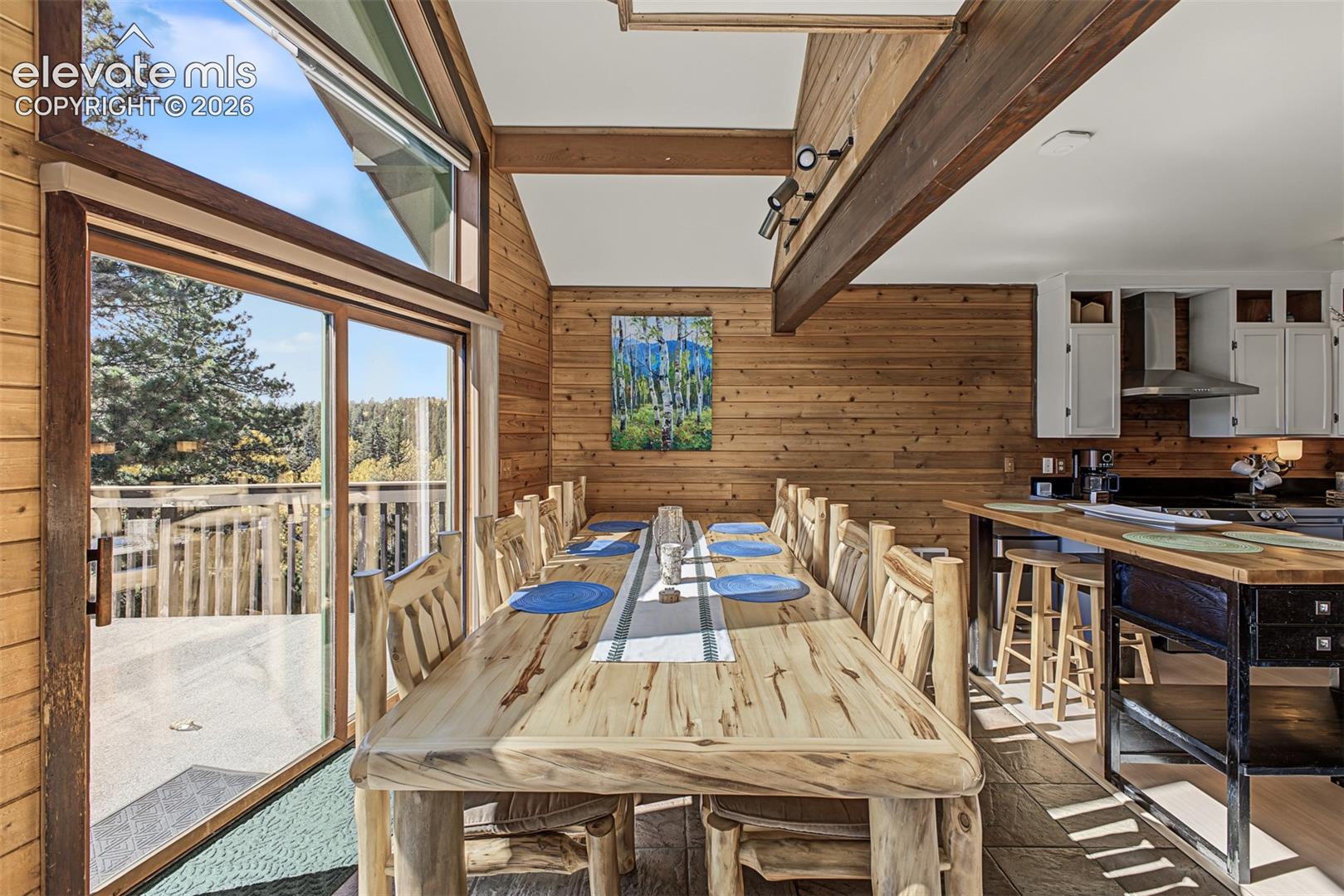 Wood-paneled interior featuring a dining area with a large wooden table, vaulted ceilings, exposed wooden beams, and expansive glass doors leading to an outdoor deck