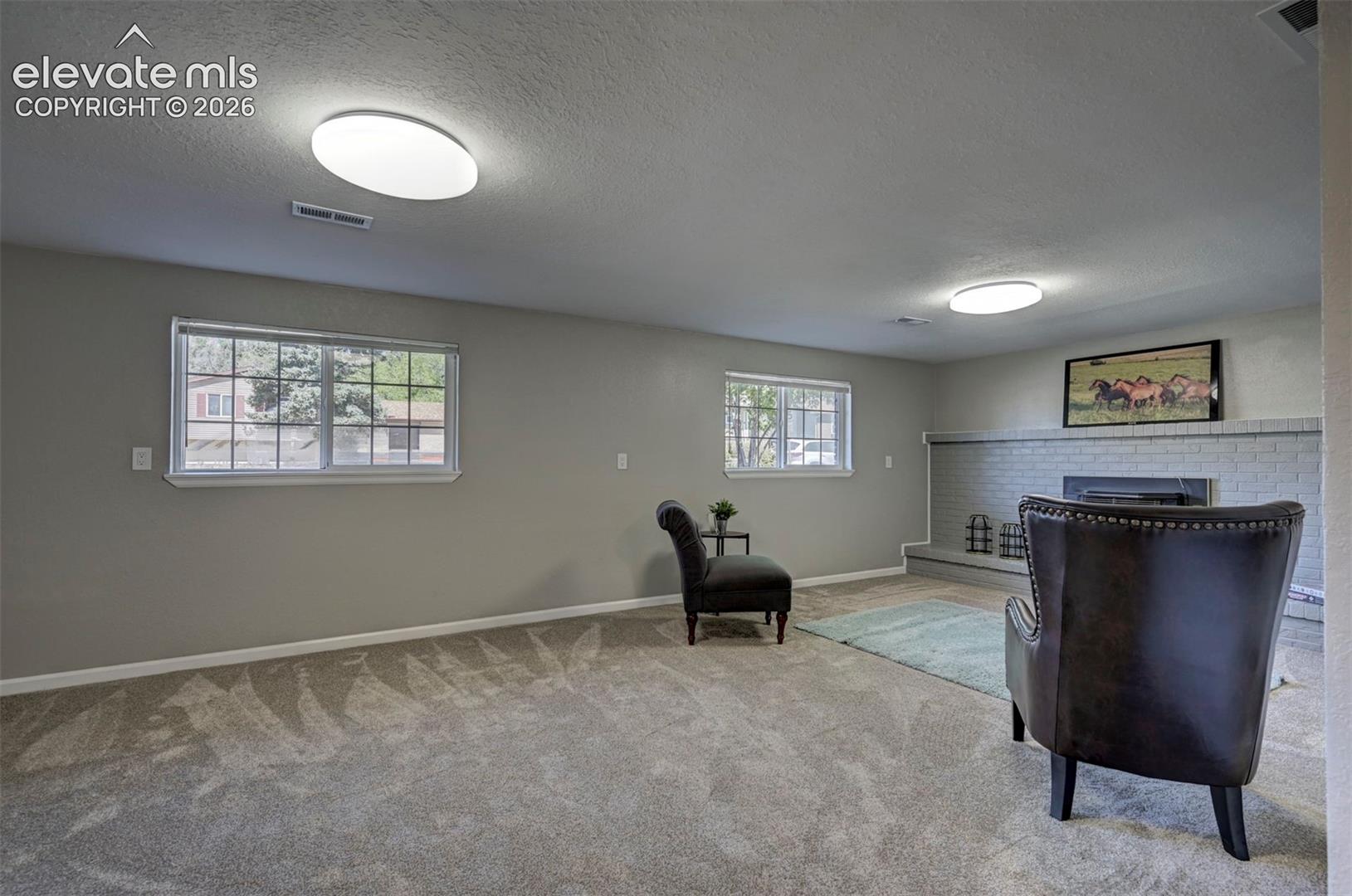 Neutral-toned interior space featuring carpeted flooring, dual ceiling-mounted light fixtures, two windows, and a painted brick fireplace surround