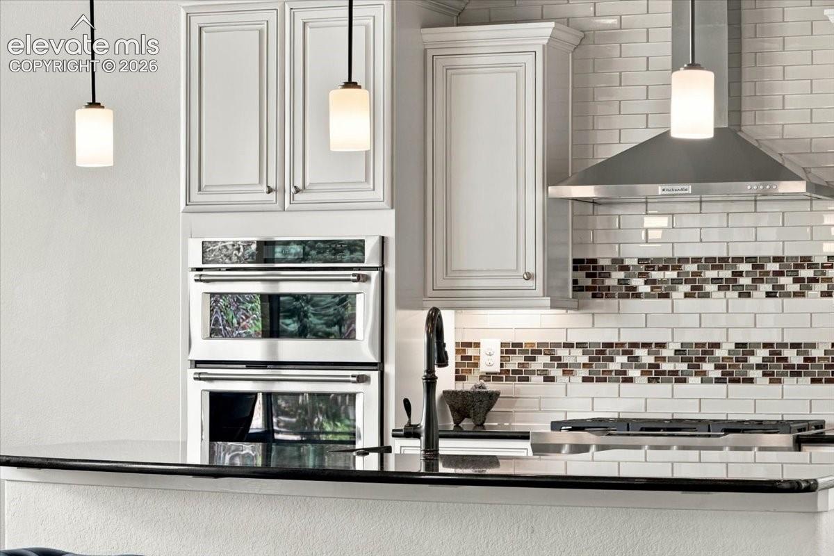 Kitchen featuring white shaker-style cabinetry, stainless steel double wall ovens, a professional-grade range hood, white subway tile backsplash with decorative mosaic inlay, and dark solid-surface countertops