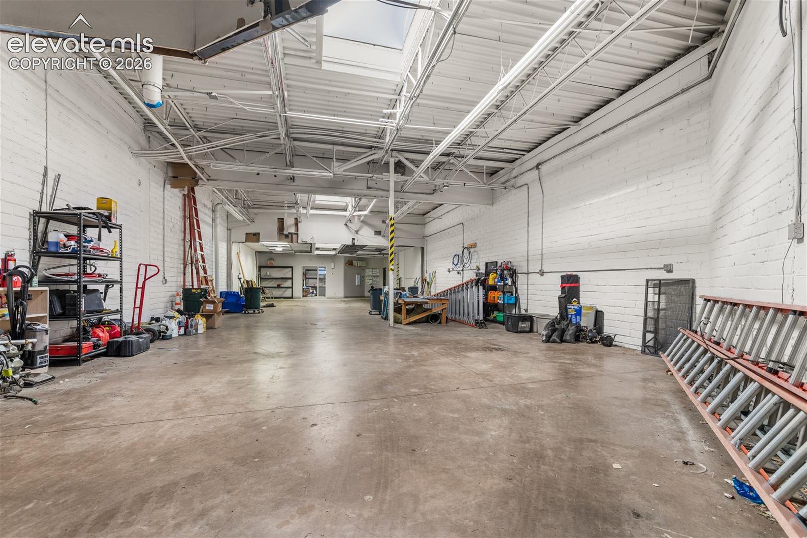 Open-concept industrial space featuring painted brick walls, exposed steel truss ceiling, concrete flooring, and multiple skylights