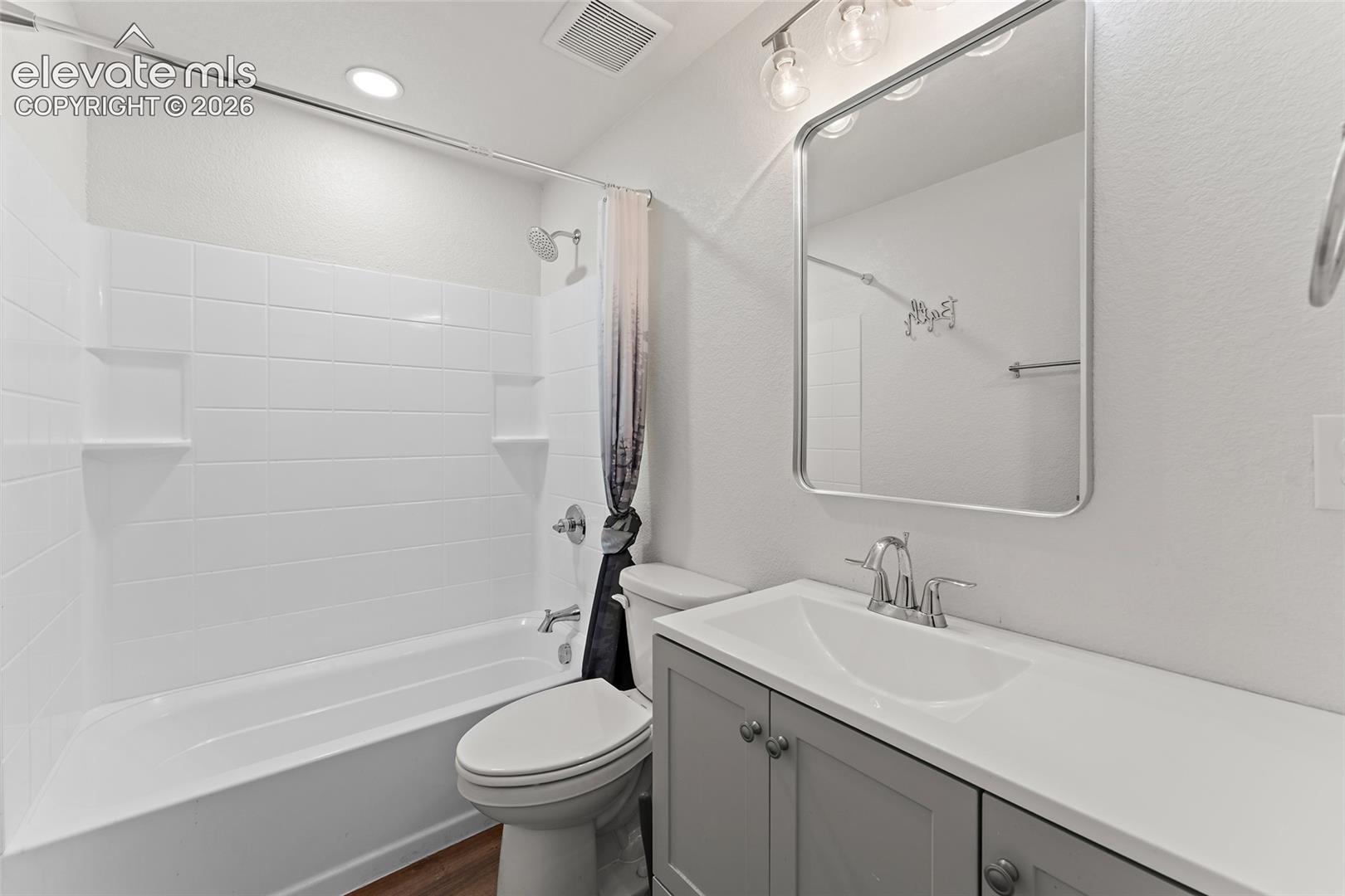 Bathroom featuring a grey vanity with a solid surface integrated sink, a rectangular mirror with rounded corners, a three-light vanity fixture, and a tub-shower combination with white tiled surround and recessed shelving
