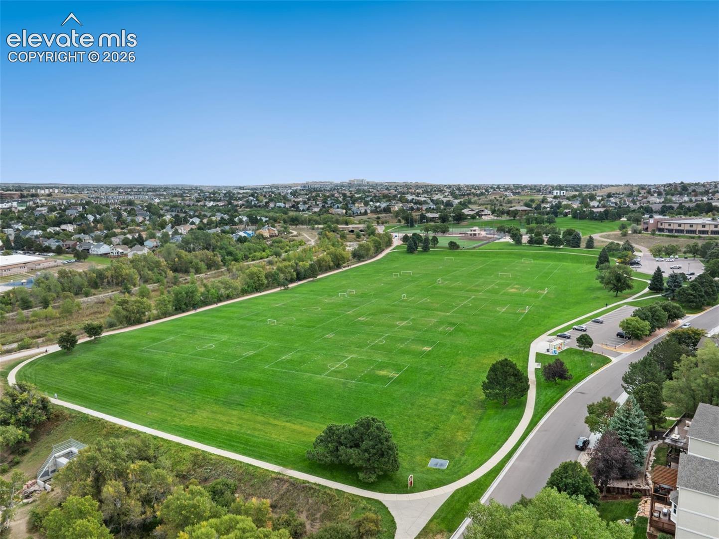 Expansive green fields with delineated sports areas, bordered by a paved pathway and mature trees, with adjacent residential structures and a parking area