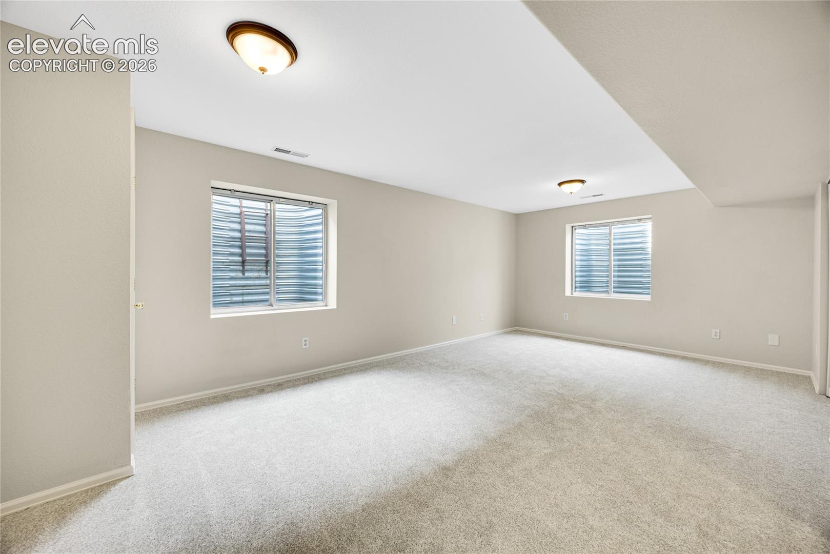 Neutral-toned room featuring wall-to-wall carpeting, two egress windows with metal window wells, and flush-mount ceiling lighting