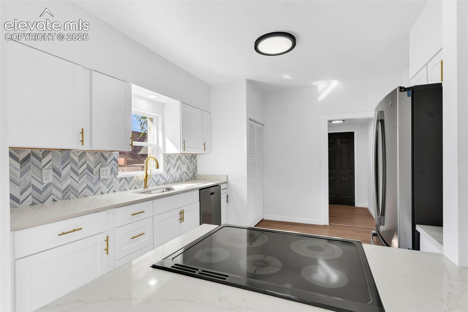 Kitchen featuring white shaker-style cabinetry, herringbone-pattern tile backsplash, quartz countertops, integrated electric cooktop, and brass-finish hardware
