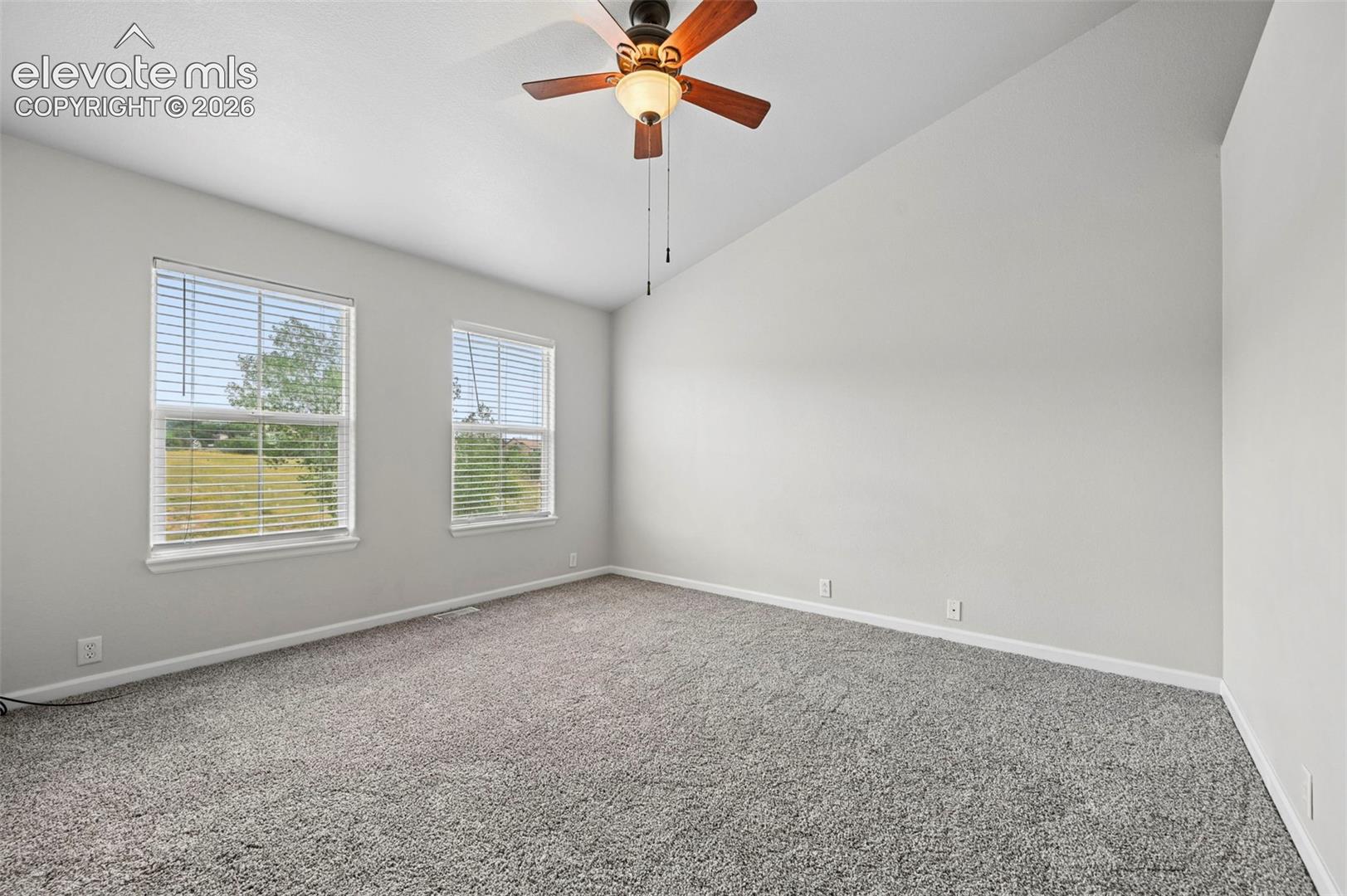 Spacious room featuring vaulted ceilings, a ceiling fan with light fixture, two windows with blinds, and light-toned carpeting