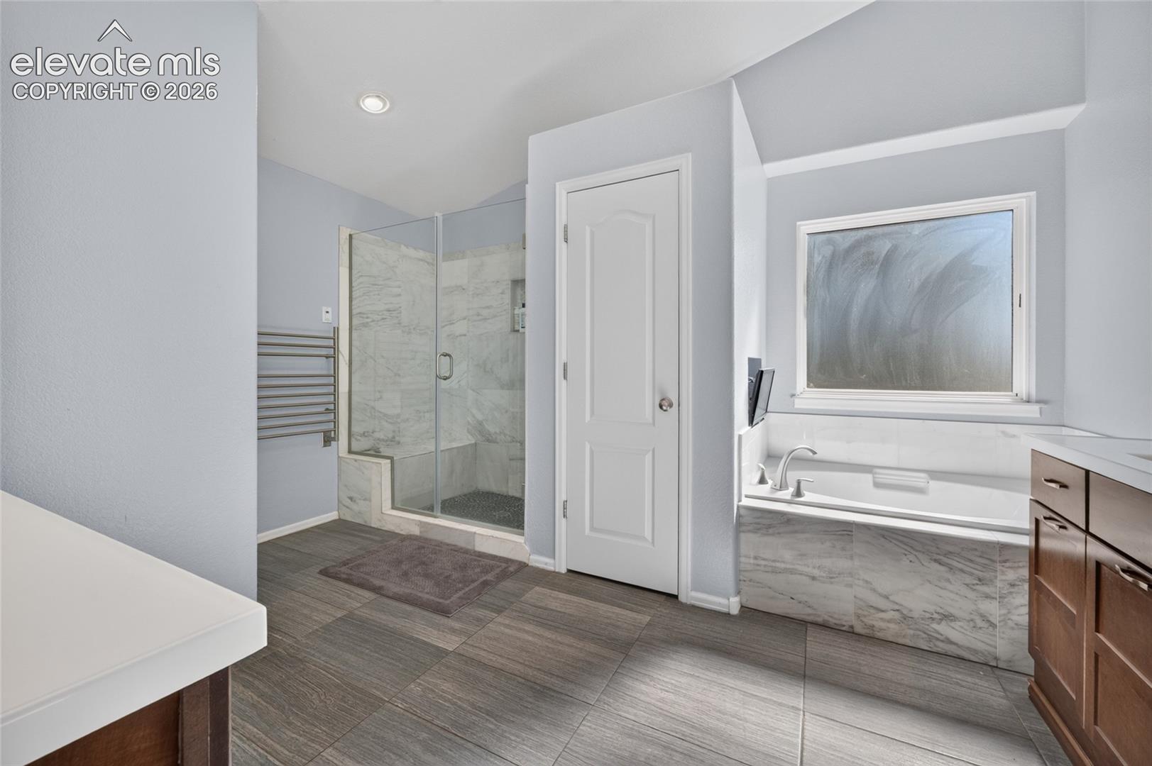Bathroom featuring a glass-enclosed walk-in shower with stone-finish tile surround, a built-in soaking tub with stone-finish tile deck, wood-finish cabinetry, and large-format tile flooring