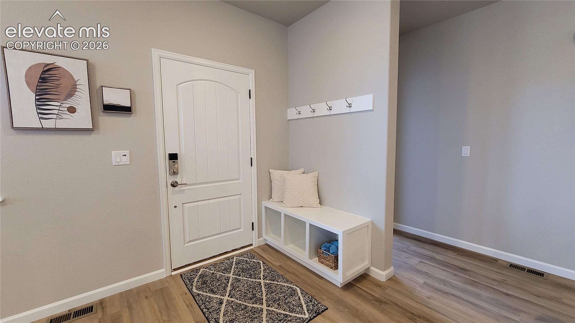Entryway featuring a white panel door with electronic deadbolt, built-in storage bench with three cubbies, wall-mounted coat hooks, and wood-finish flooring