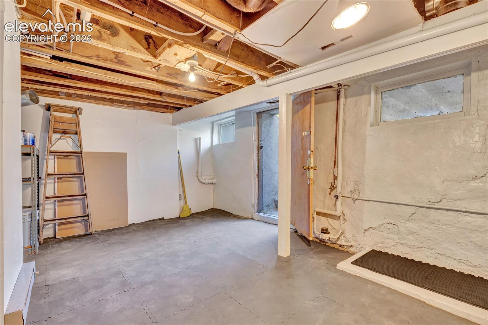 Lower level storage area featuring exposed wood ceiling joists, painted concrete walls, concrete flooring, and an exterior access door
