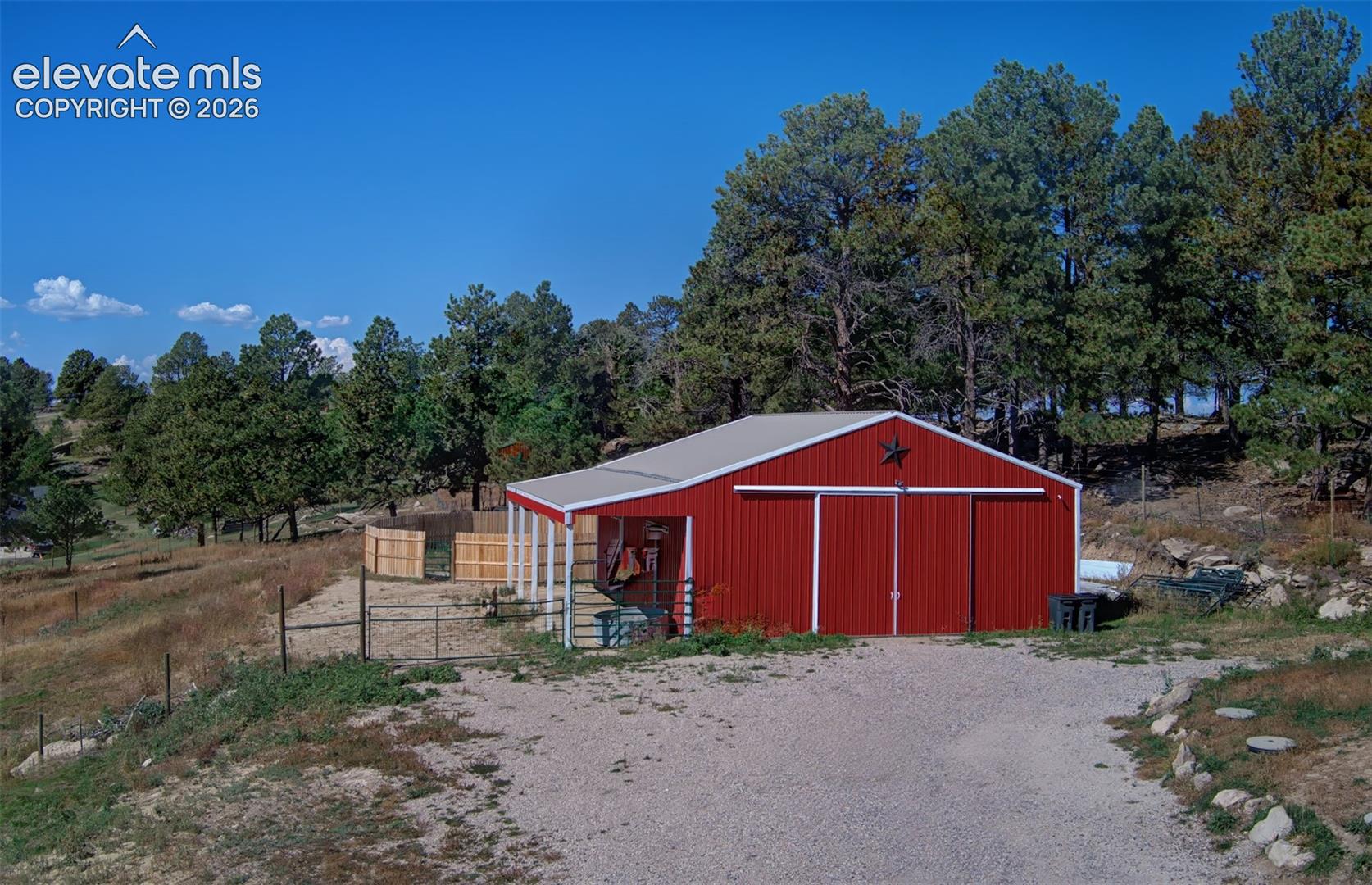 Barn featuring red metal siding, a gabled roof, sliding barn doors, and an attached side-porch, situated on a lot with perimeter fencing and mature trees