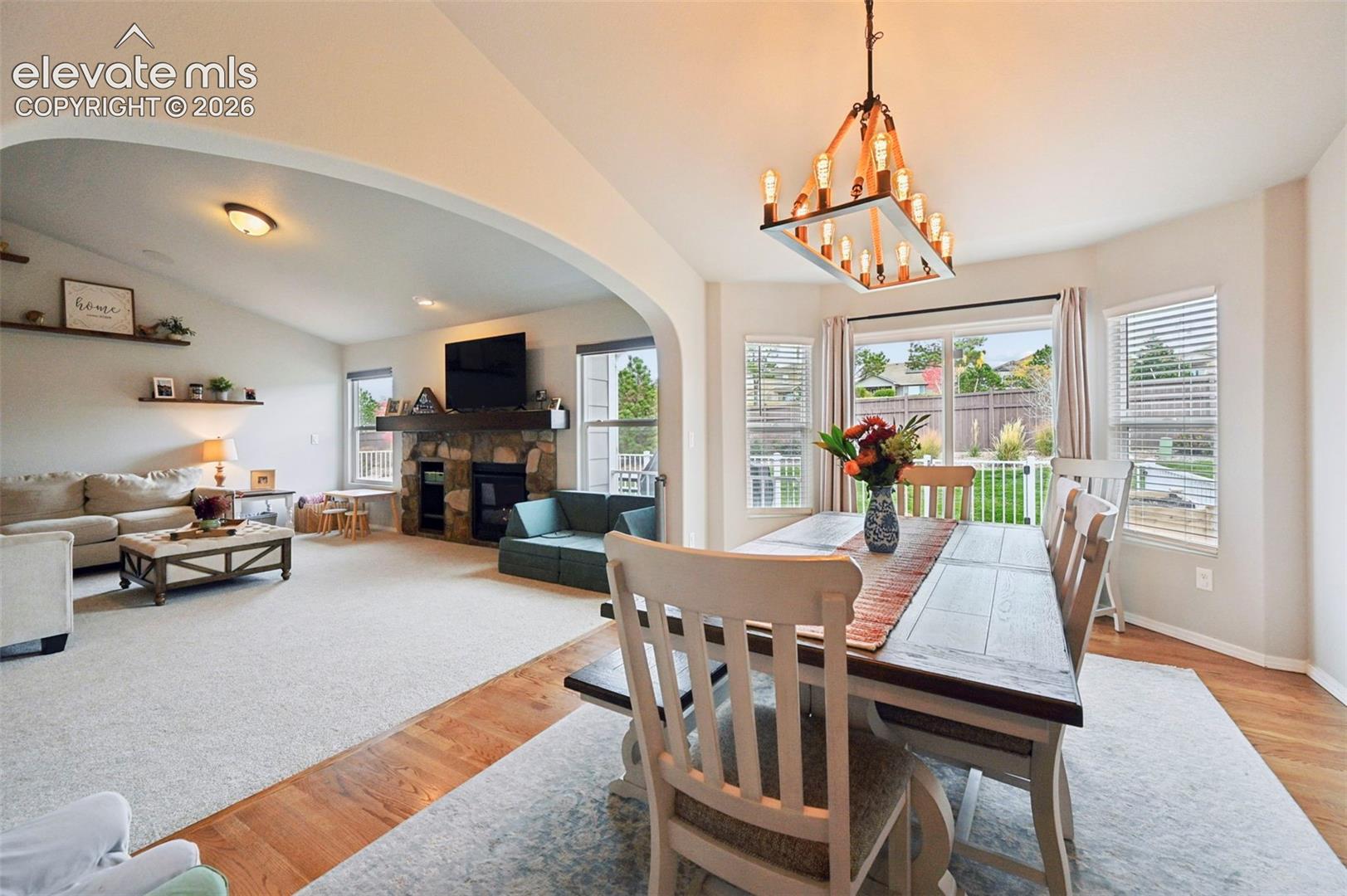 Vaulted ceilings with an arched entryway, stone-faced fireplace with a timber mantel, rectangular lantern-style chandelier, and sliding glass patio doors