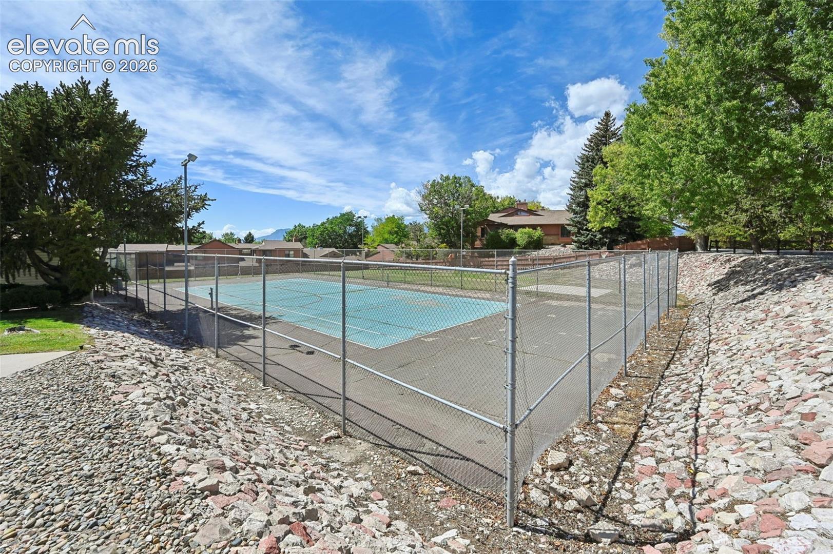 Fenced outdoor court with a hard surface, featuring overhead lighting and surrounding mature trees