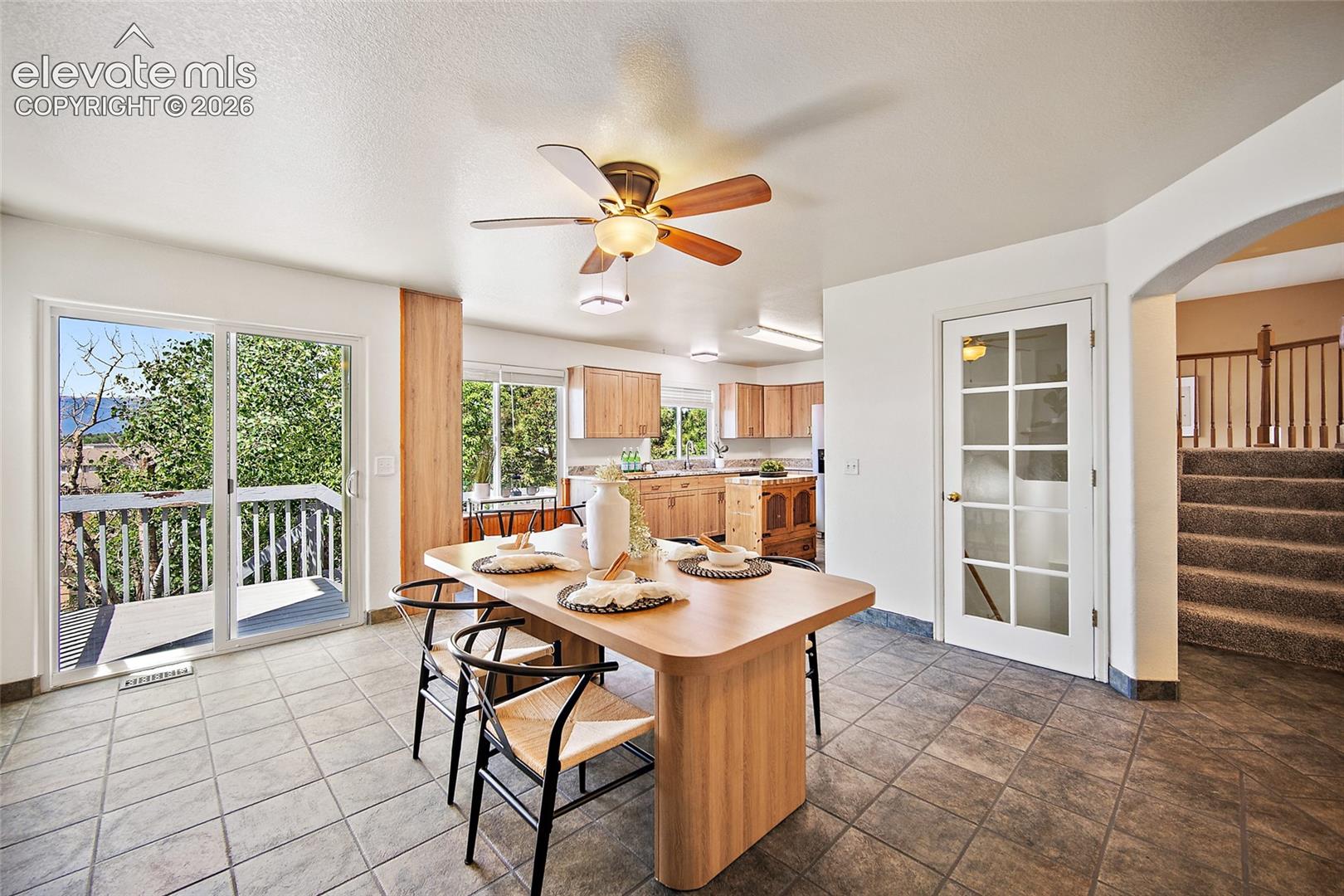 Tile flooring extending from the dining area into the kitchen, featuring sliding glass doors to a private deck, a ceiling fan with integrated lighting, a white French pane door, and an arched entry to a carpeted staircase