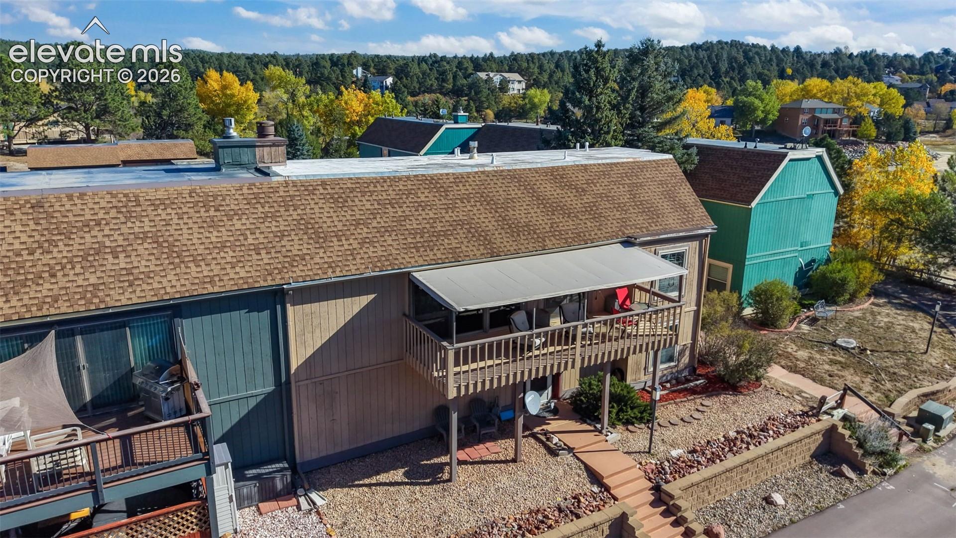 Multi-level exterior featuring a shingle roof, raised wood deck with overhead canopy, wood-paneled siding, and stone hardscaping