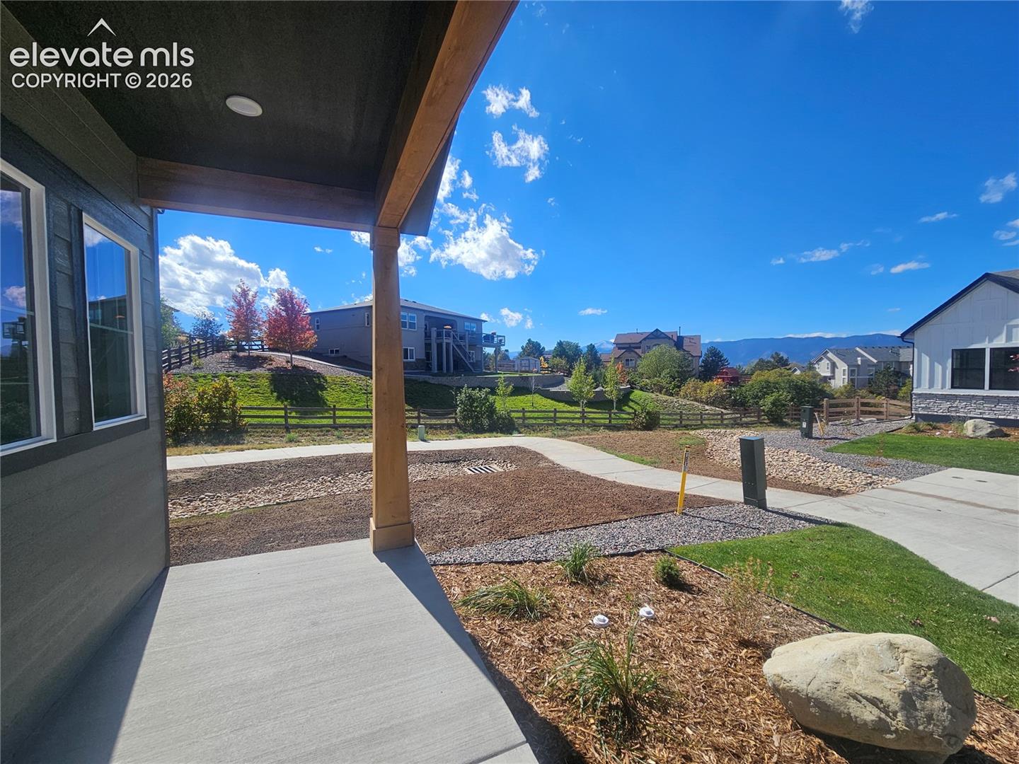 Covered patio featuring wood-wrapped support posts, concrete flooring, exterior siding with horizontal texture, and xeriscaped grounds with gravel beds and decorative boulders
