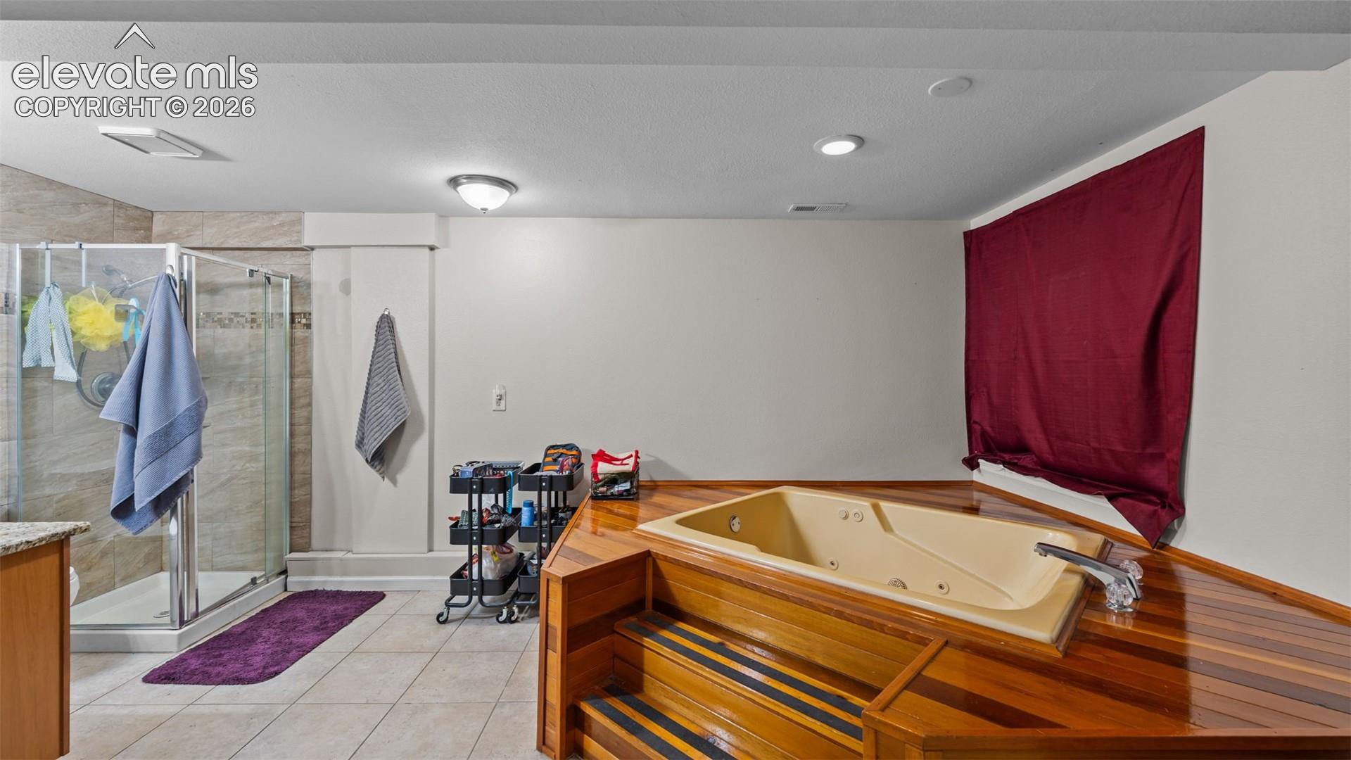 Soaking tub featuring wood-finish decking and tiered steps, tiled walk-in shower with glass enclosure, recessed lighting, and tile flooring