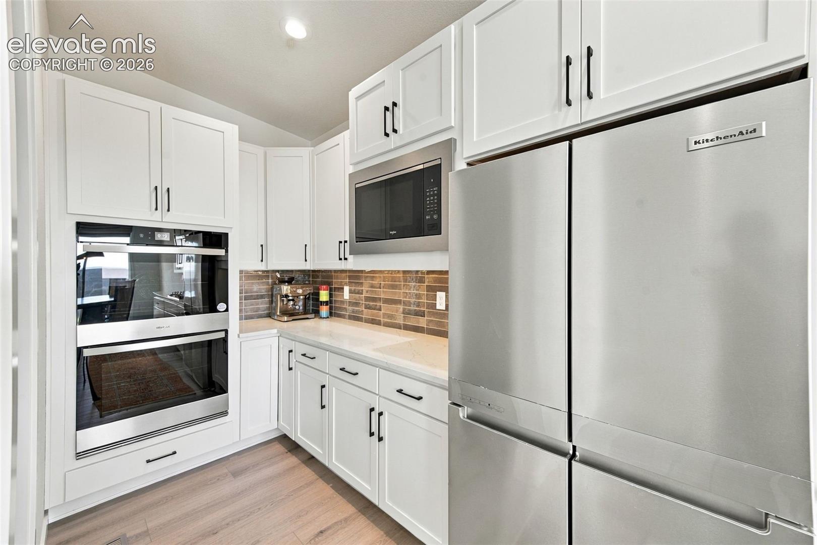 Kitchen featuring white shaker-style cabinetry, stainless steel appliances including a double wall oven and refrigerator, textured tile backsplash, and light wood-finish flooring
