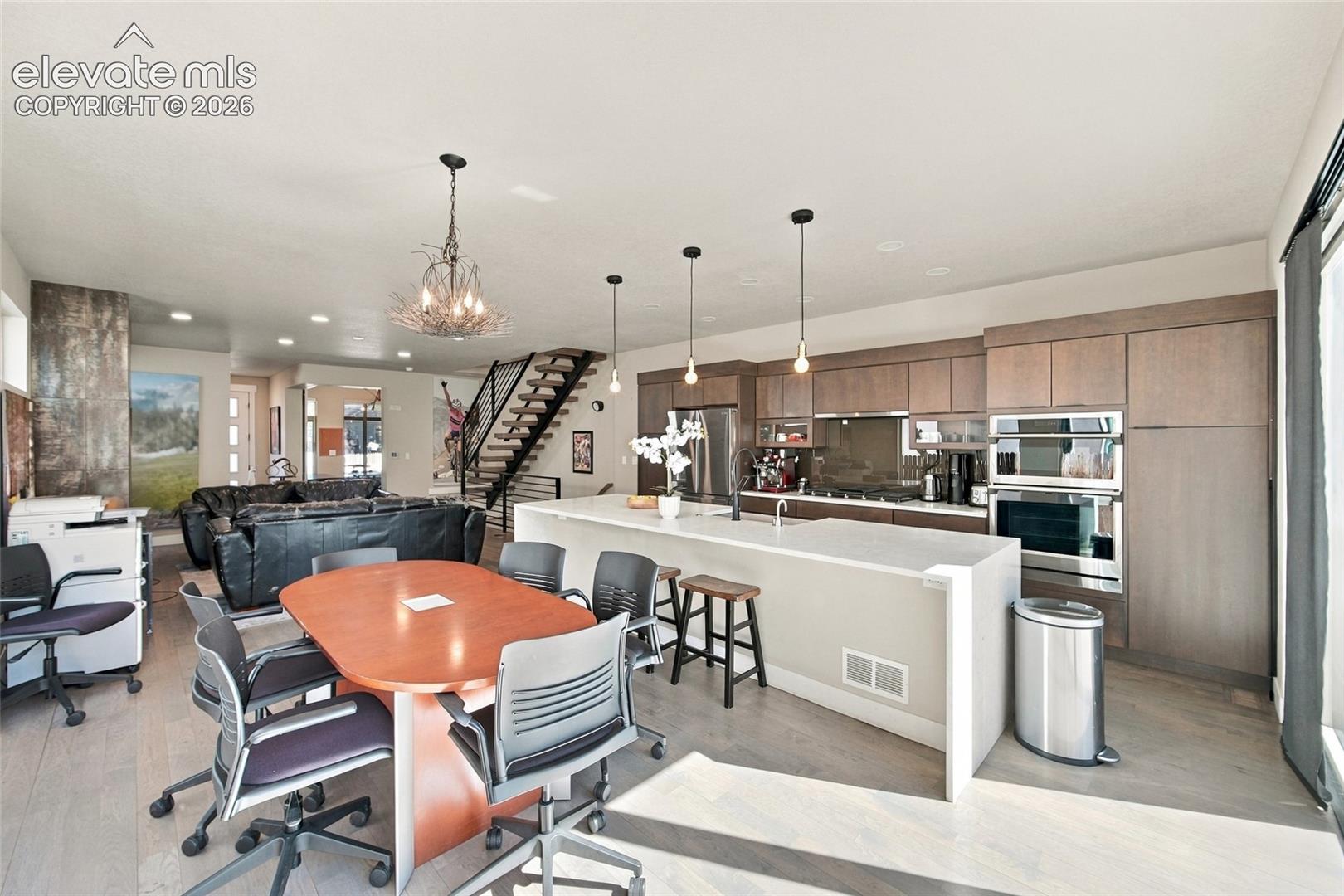 Open-concept kitchen featuring flat-panel cabinetry, a central island with a waterfall countertop, stainless steel wall ovens, wood flooring, and a floating staircase with metal railing