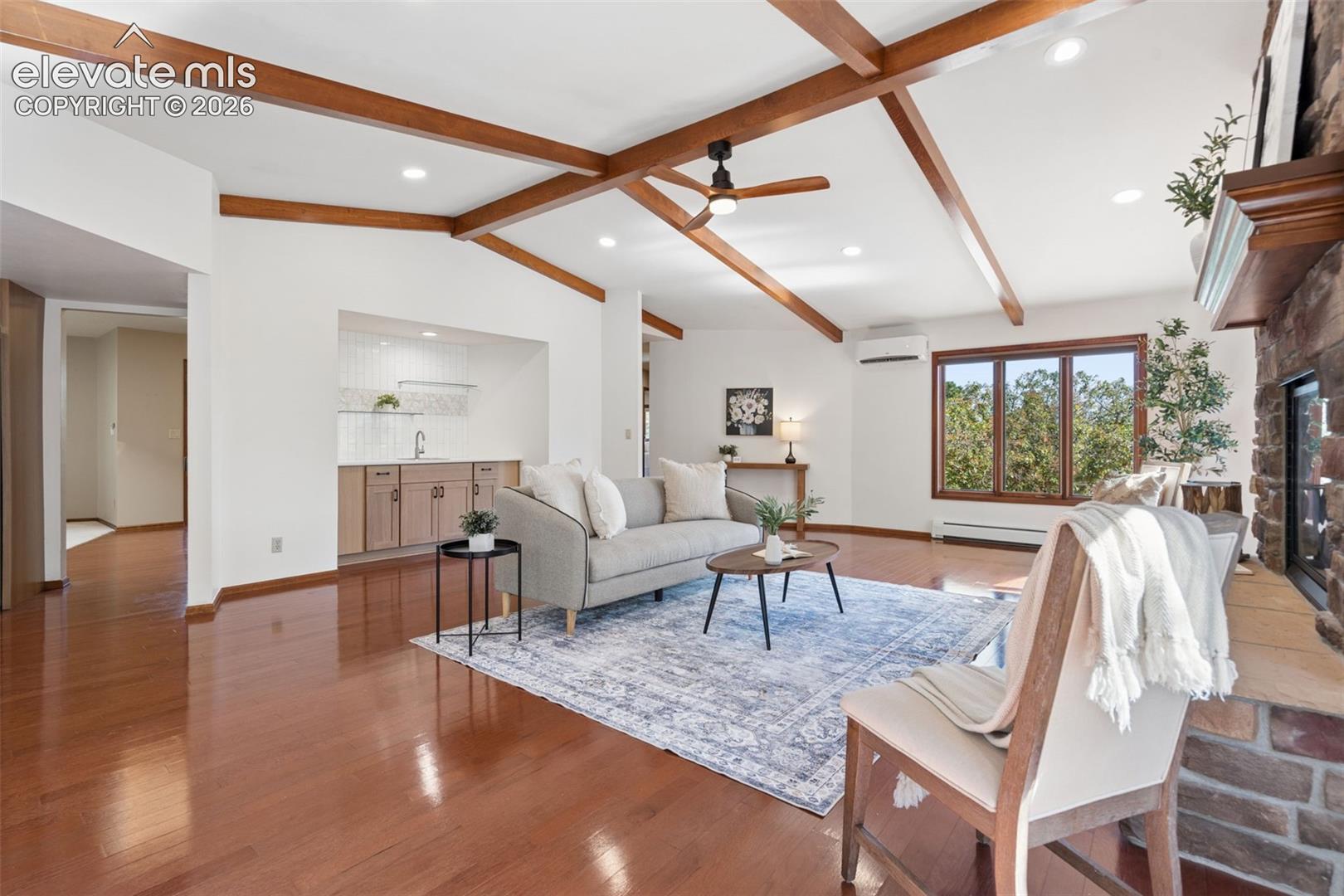 Vaulted ceiling with exposed wood beams, wood-finish flooring, stone-clad fireplace, built-in wet bar, and large picture windows