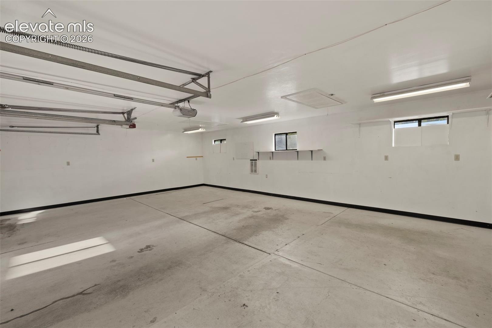 Spacious garage with concrete flooring, multiple windows, built-in shelving, and overhead fluorescent lighting