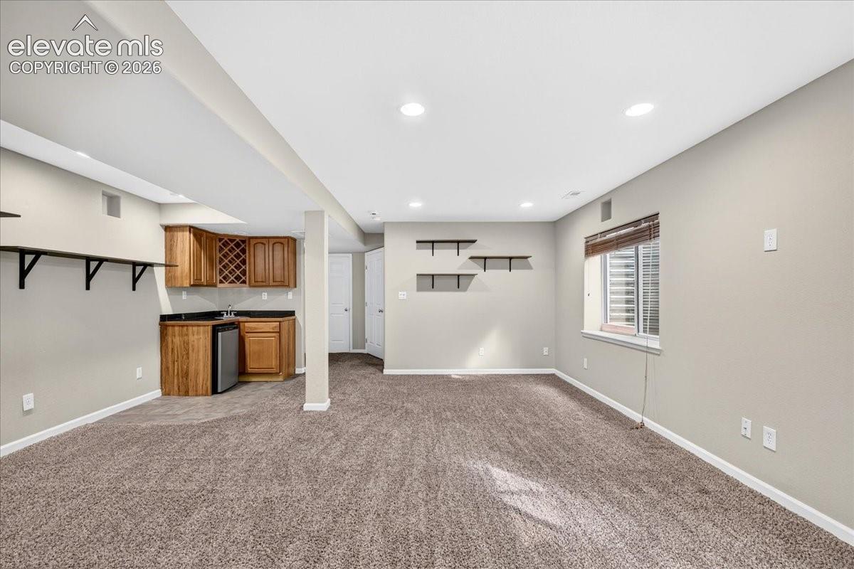 Recessed lighting illuminates the carpeted living area, featuring a built-in wet bar with wood cabinetry and a stainless steel compact refrigerator