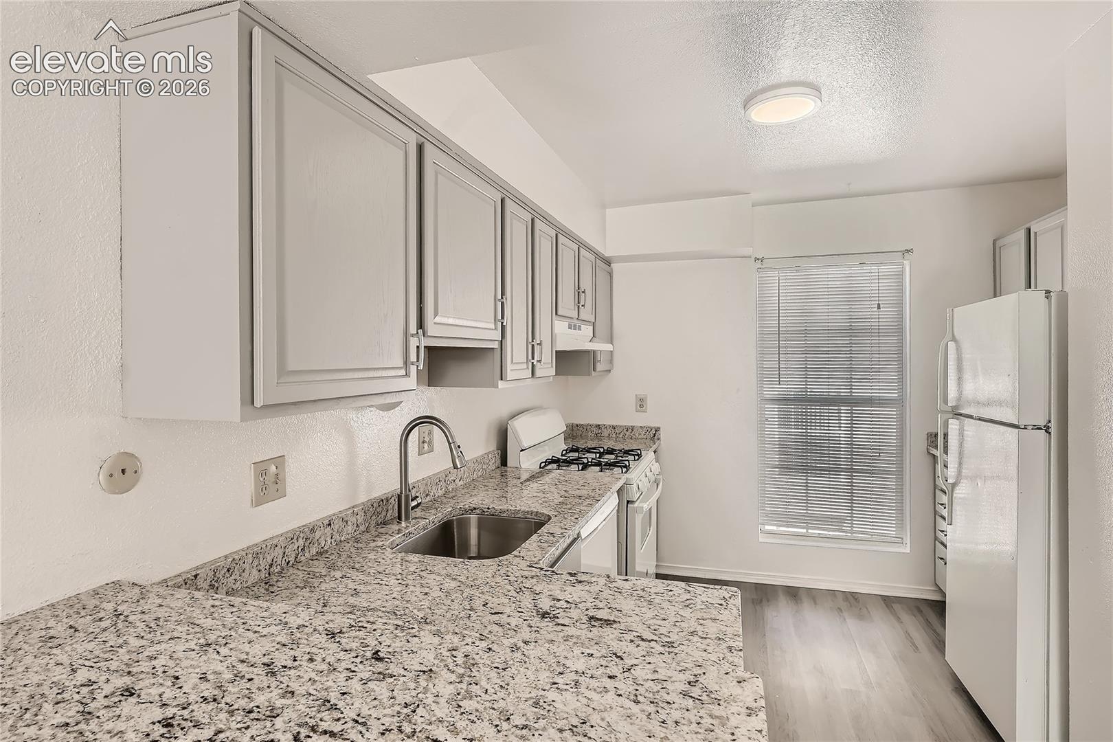 Kitchen featuring light-toned cabinetry, stone-surface countertops, an undermount stainless steel sink with a high-arch faucet, and wood-finish flooring