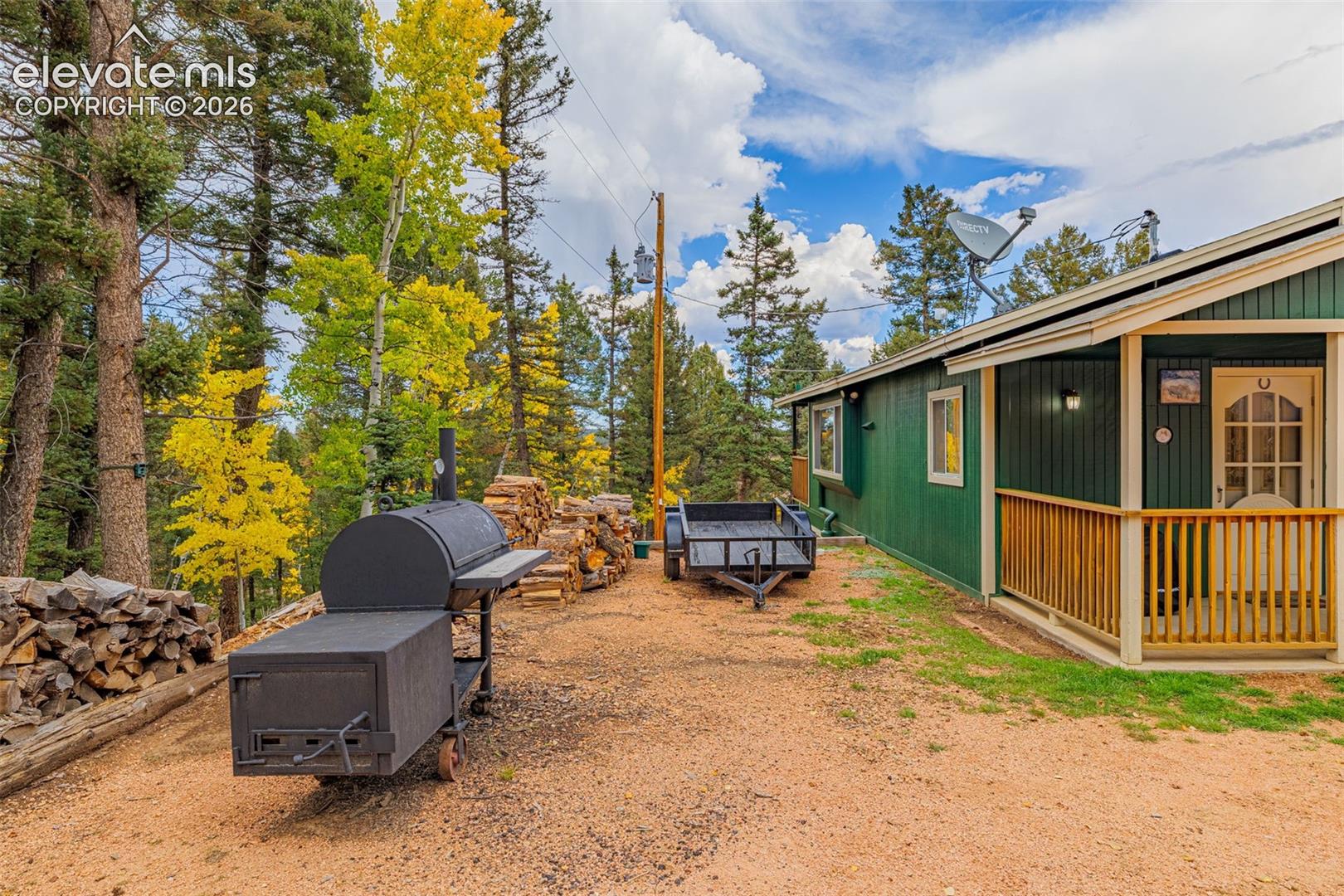 Exterior of a residence featuring vertical green siding, a covered porch with a wood-railing, and a gravel grounds area surrounded by mature pine and aspen trees
