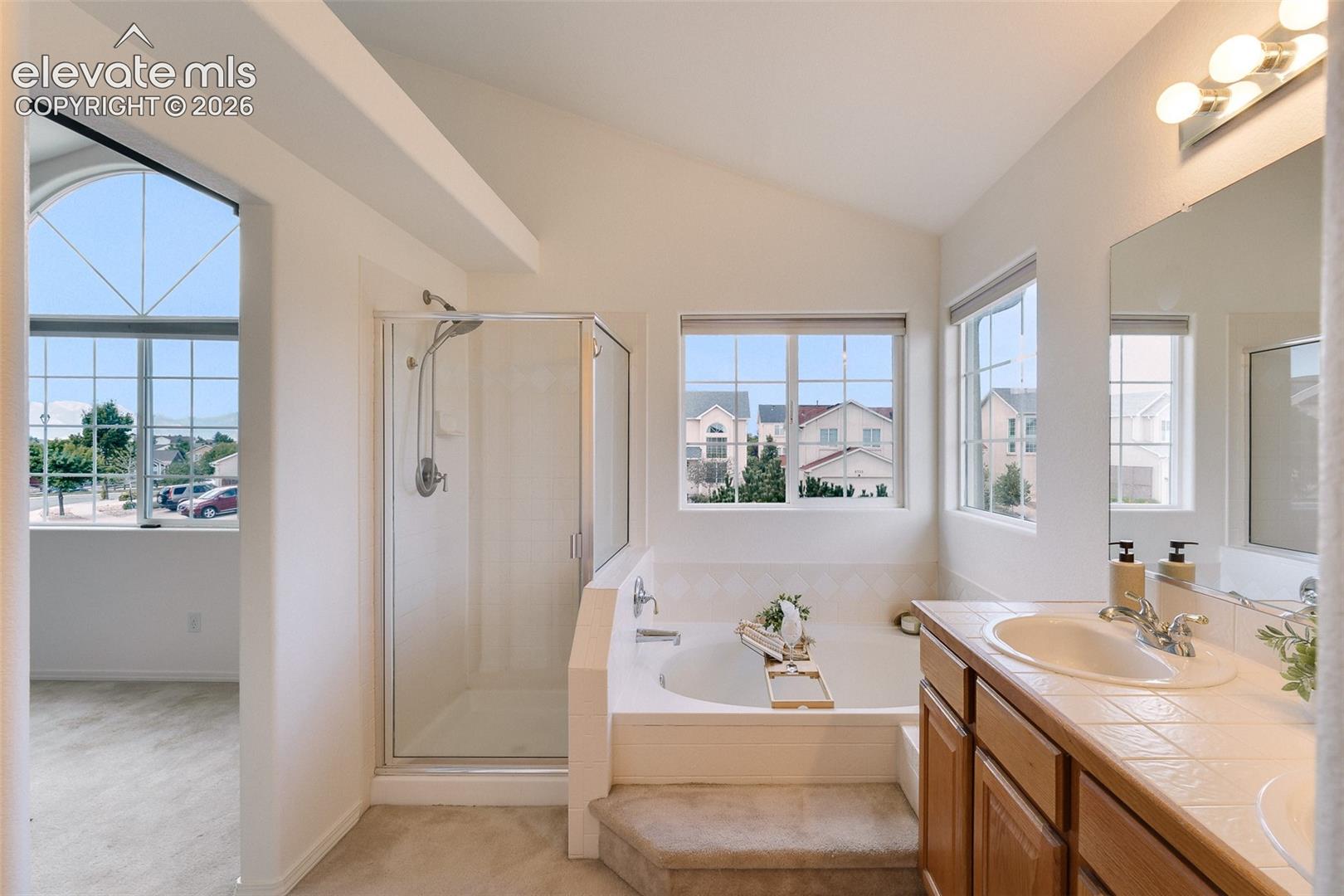 Vaulted ceiling, glass-enclosed shower, soaking tub with tiled surround, wood vanity with integrated sink, and large mirror