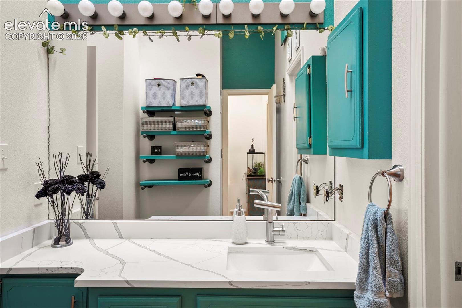 Bathroom vanity featuring a stone-composite countertop with integrated rectangular basin, single-handle brushed nickel faucet, wall-mounted theater-style light fixture, large plate glass mirror, and built-in shelving