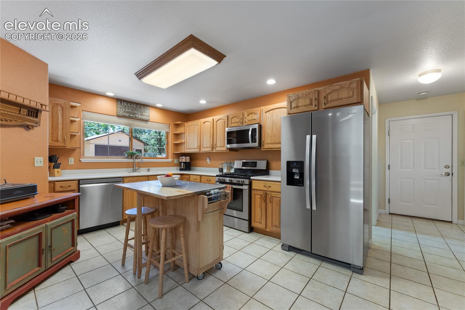 Kitchen featuring oak cabinetry, stainless steel appliances, a center island with a wood-finish base, tile flooring, and a large window above the sink