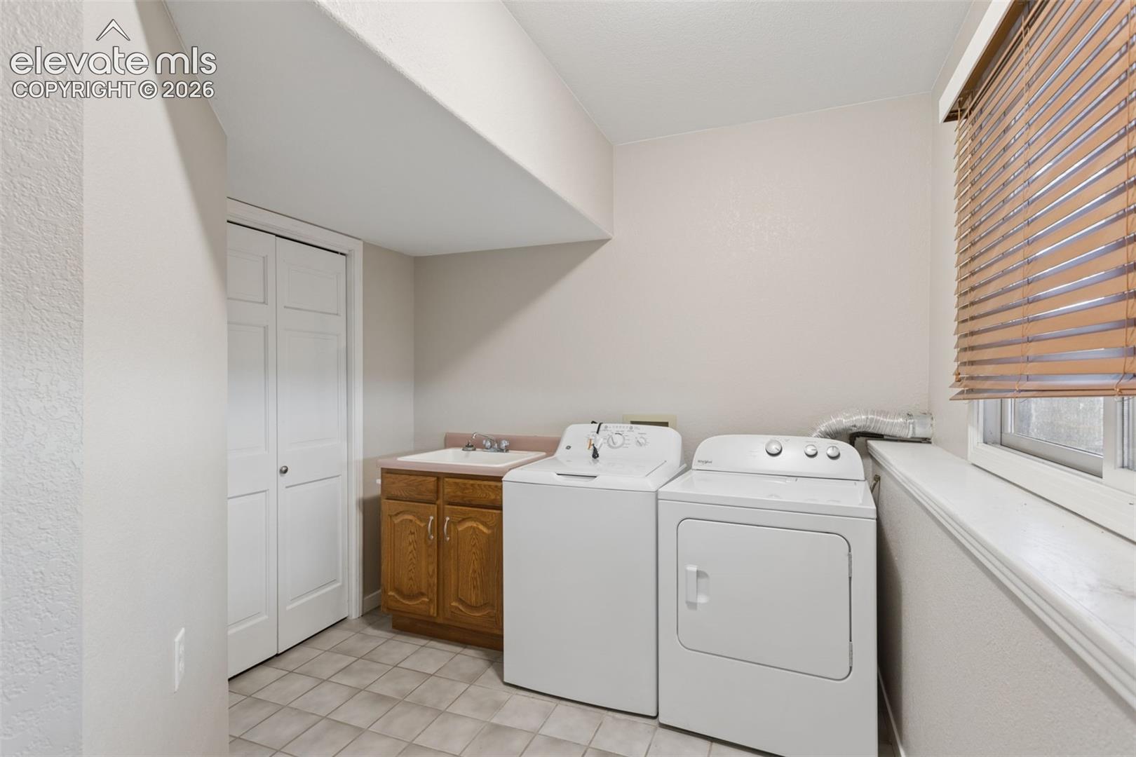 Laundry area featuring a utility sink with wood-finish cabinetry, tile flooring, and a window with blinds