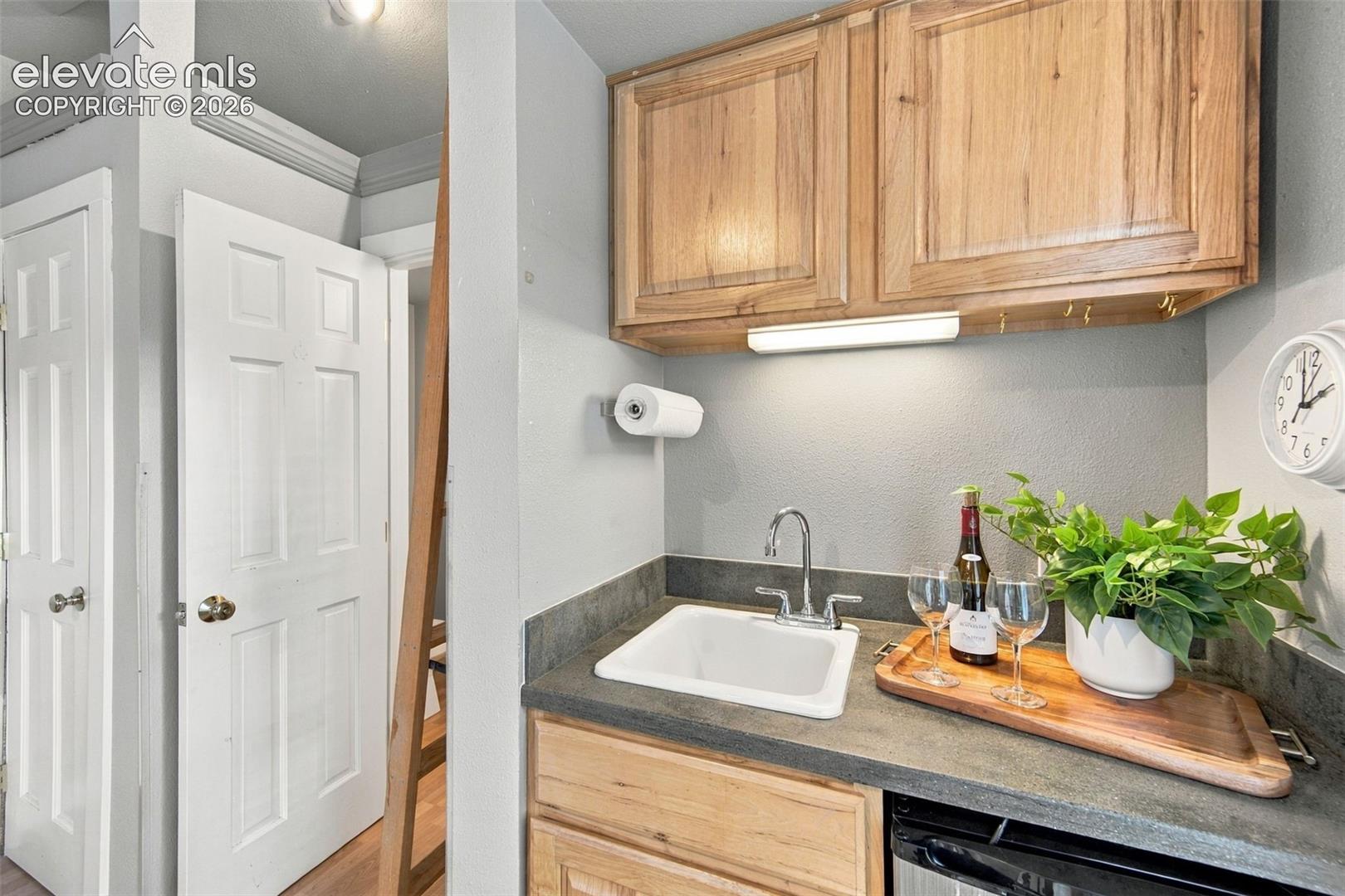 Wet bar featuring natural-finish wood cabinetry, solid-surface countertop, under-cabinet lighting, and a drop-in sink with a chrome faucet