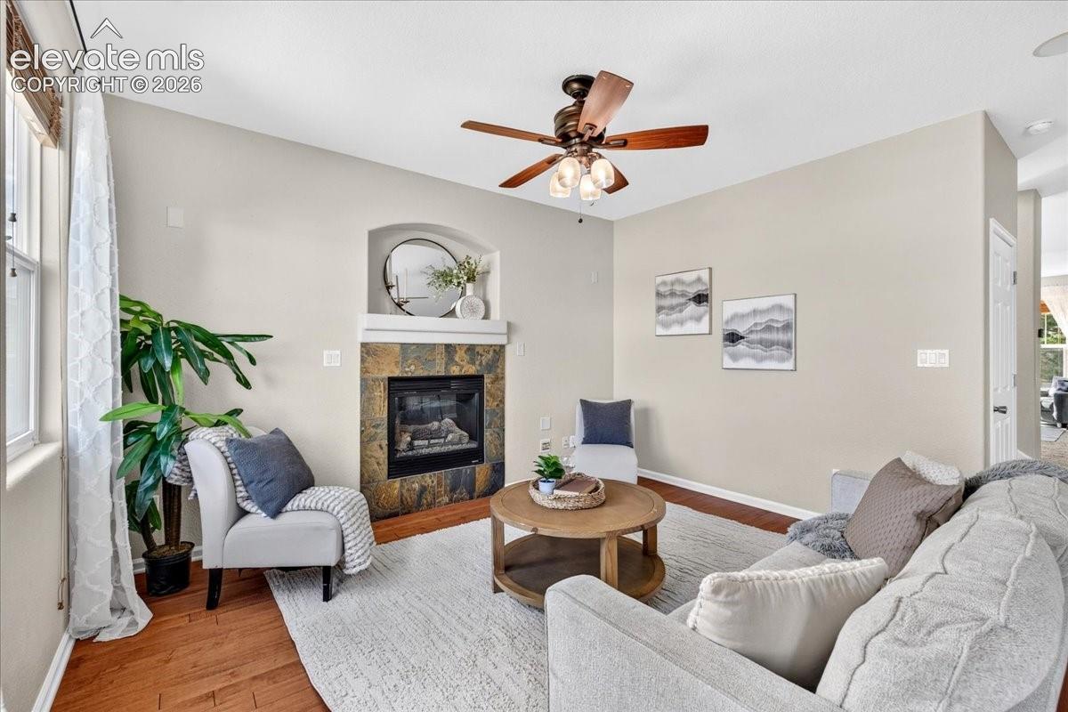 Fireplace with stone surround and mantle, vaulted ceiling, ceiling fan with integrated lighting, window with blinds, and wood-finish flooring